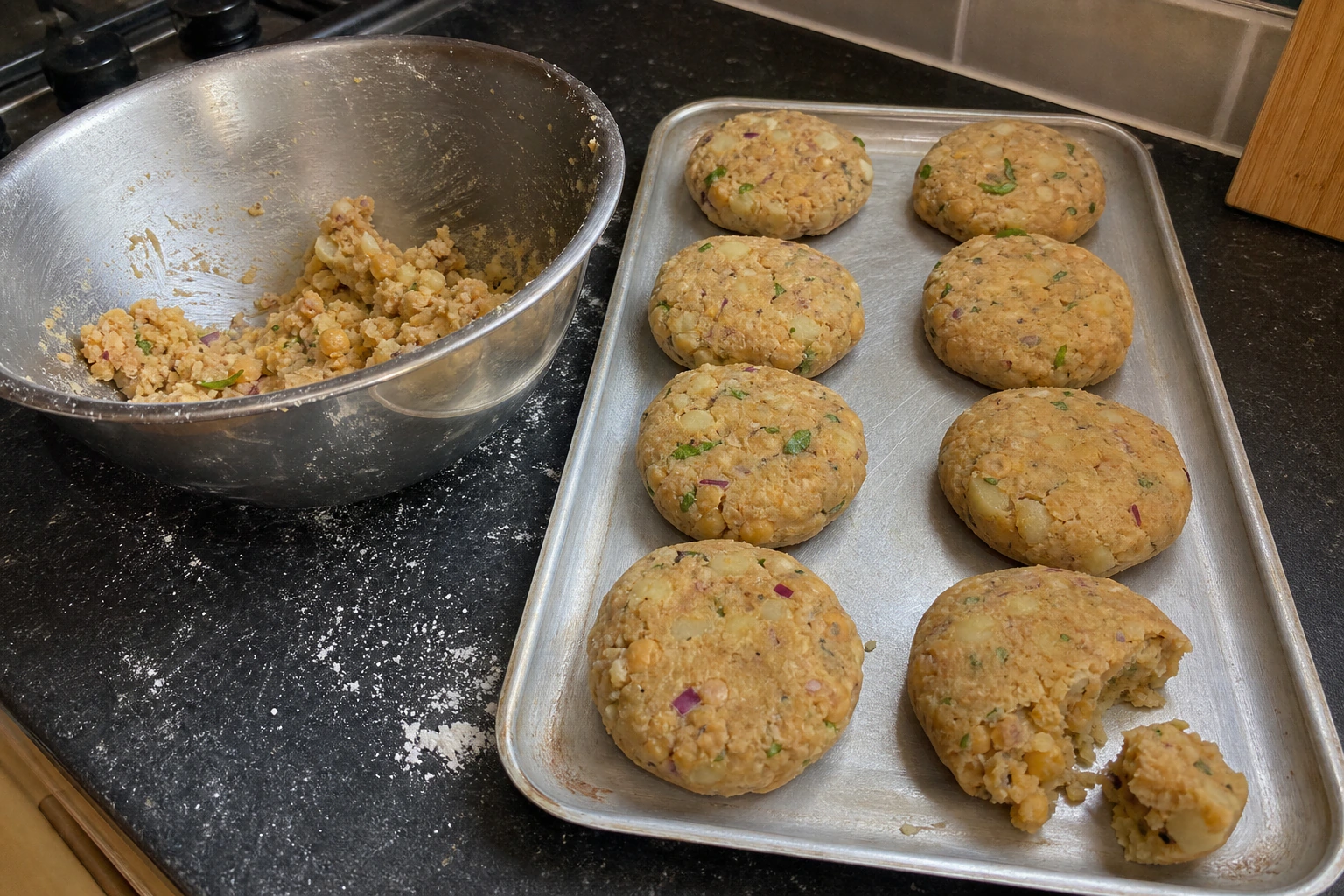 Eight thick evenly shaped aloo tikki patties on a tray with one broken open to show the potato texture