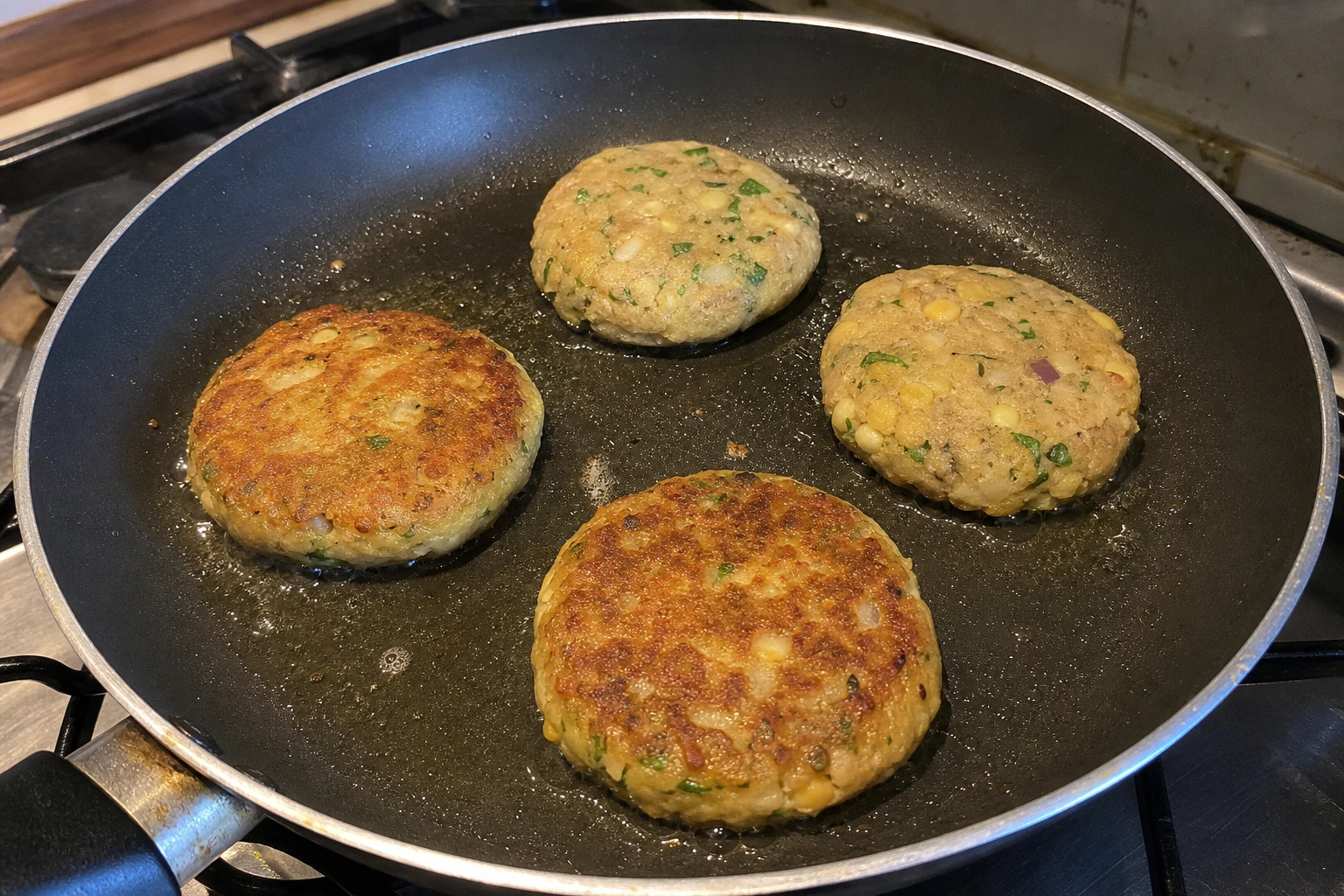 Aloo tikkis frying in a shallow pan with two deeply golden patties and two still pale before flipping