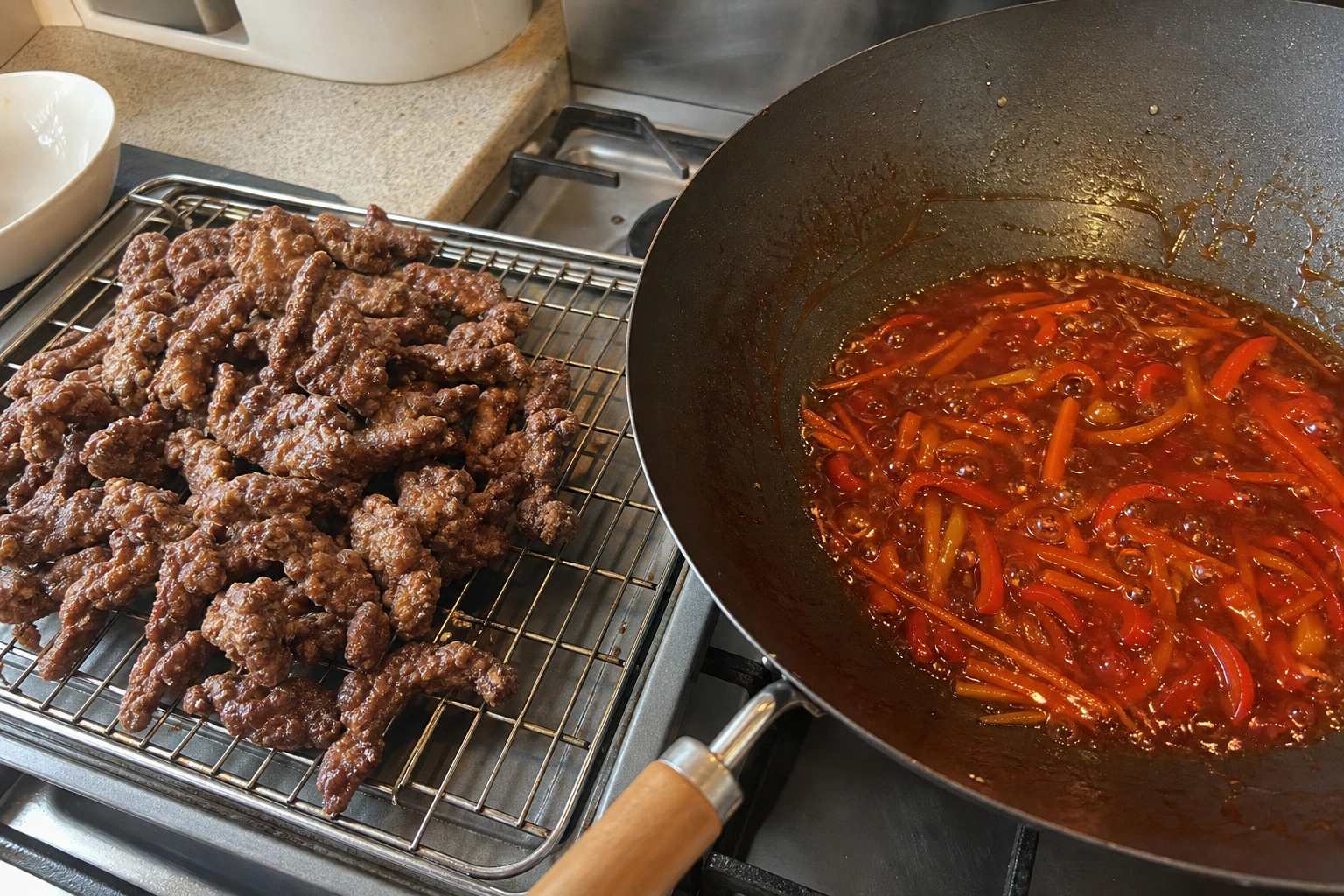 Crispy beef resting on a wire rack beside a wok of bubbling sticky chilli sauce before the final toss