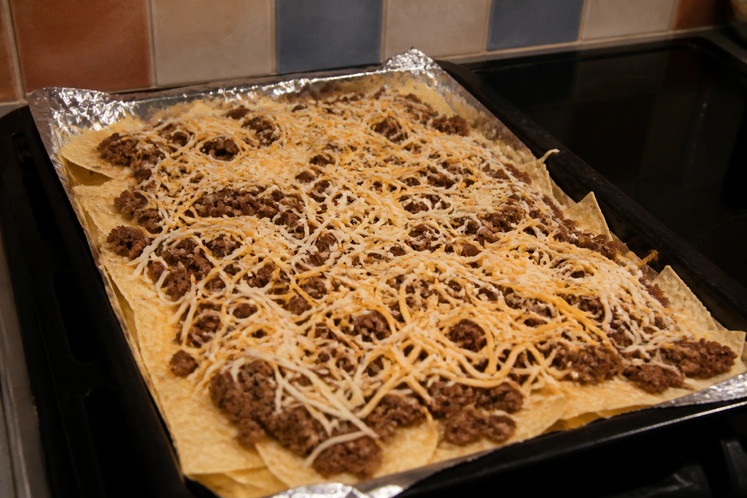 A wide tray of nachos being built in two even layers of tortilla chips beef and cheese before baking