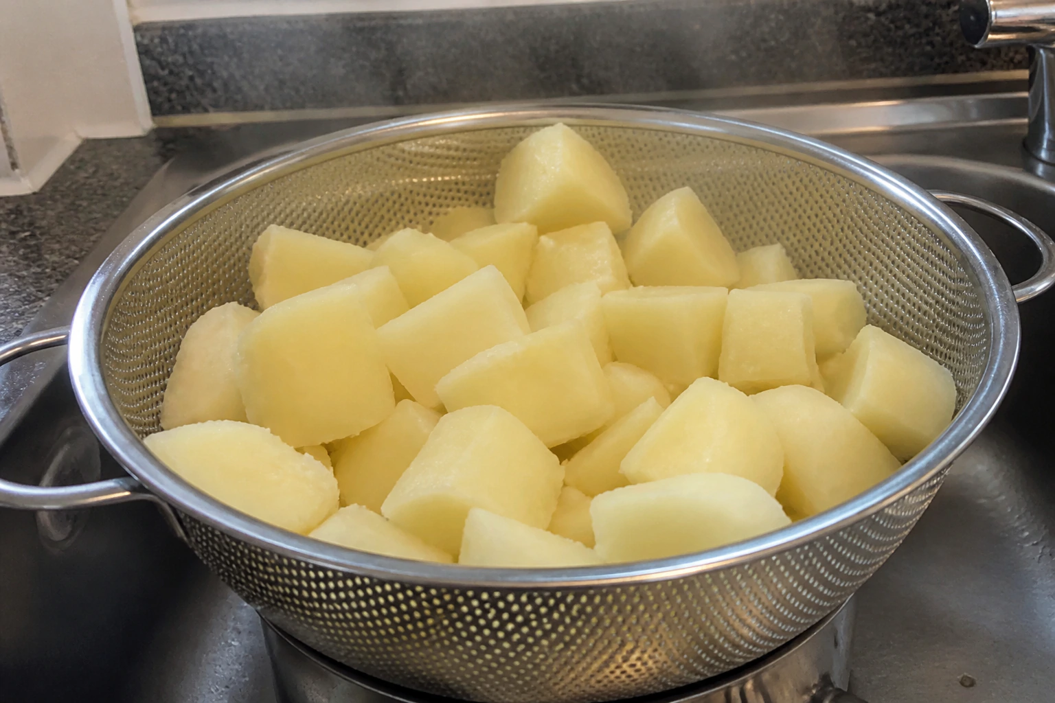 Parboiled potato chunks spread out to steam dry with chalky dry surfaces in a home kitchen colander
