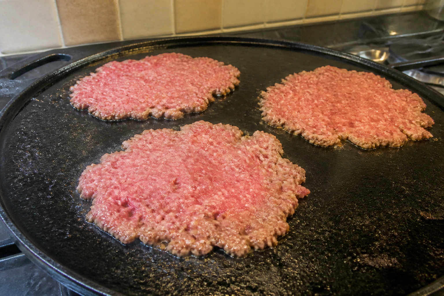 A loose ball of beef being pressed into a very thin smash burger patty on a hot cast-iron pan