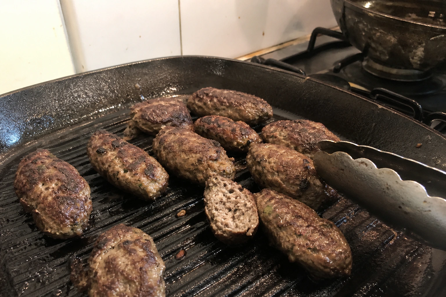 Uneven hand-shaped lamb koftas on a used griddle with one cut centre showing fully cooked mince