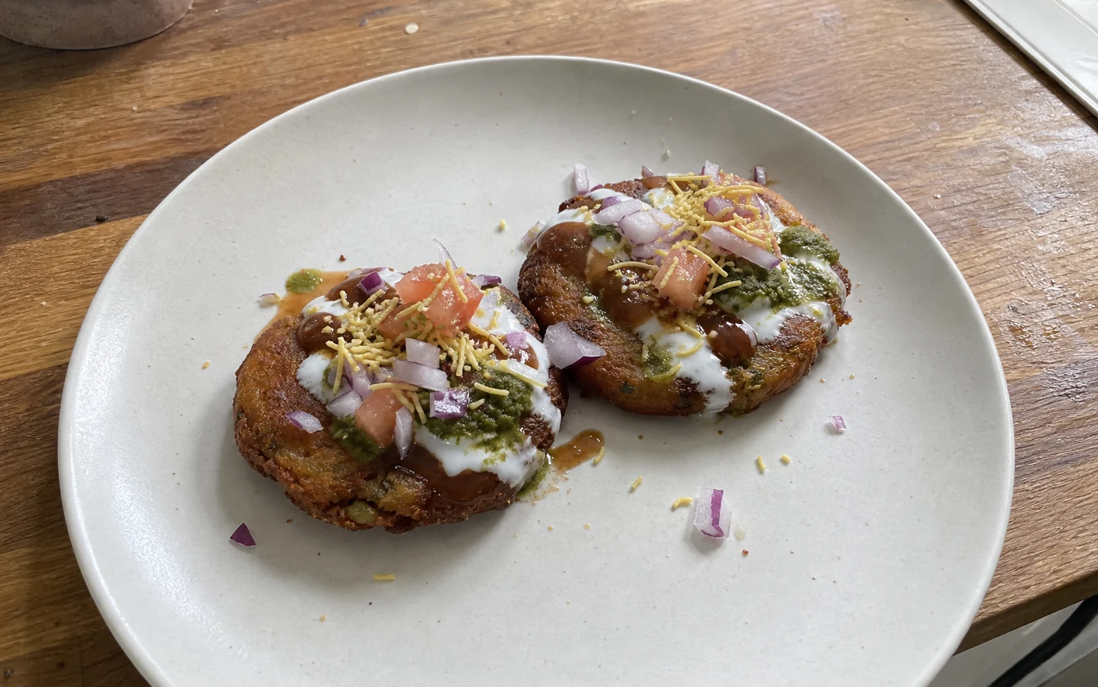 Two crisp aloo tikkis with restrained yoghurt, tamarind, mint chutney, onion, tomato and sev on a kitchen table