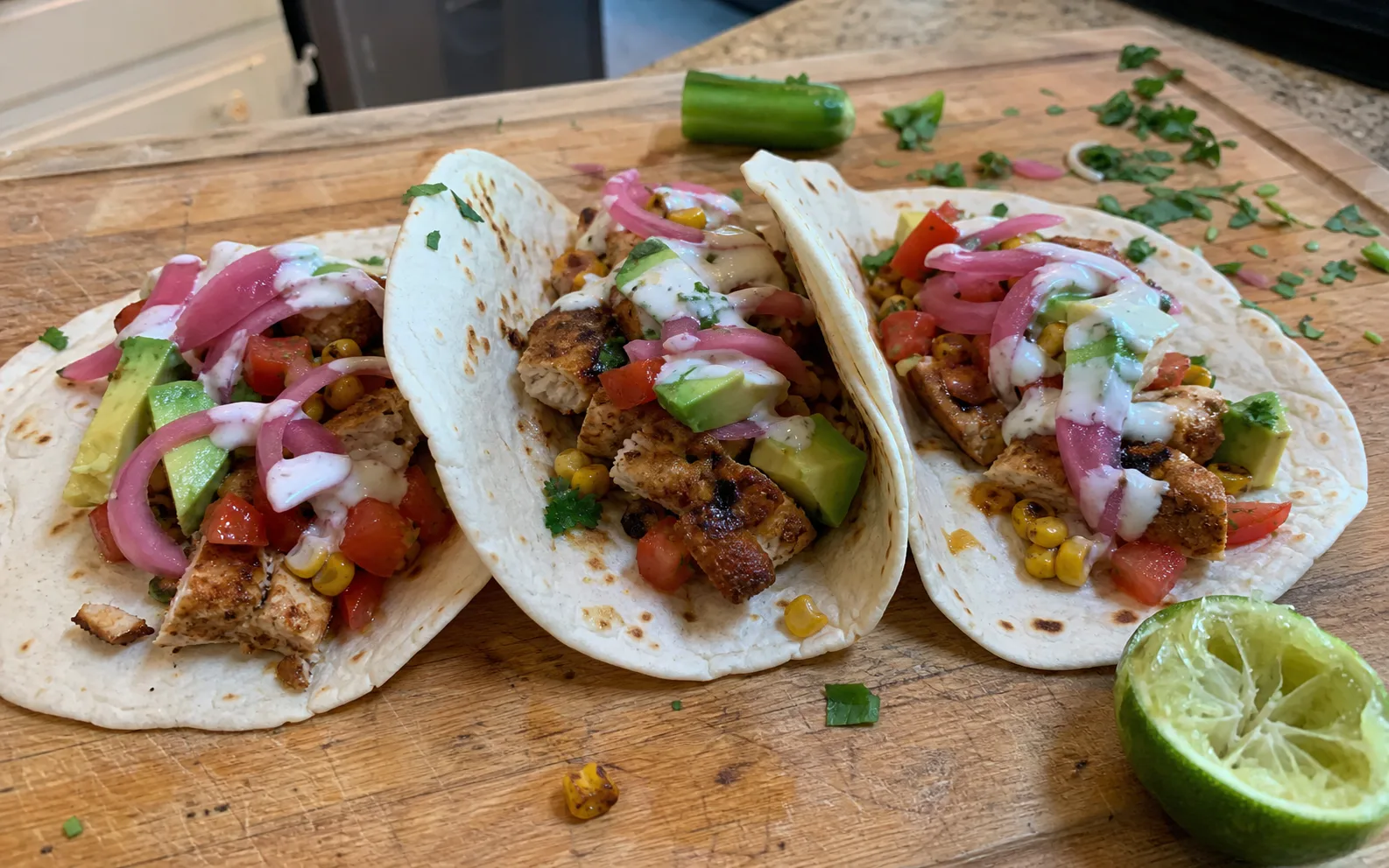 Three homemade chicken tacos with avocado, corn, salsa, pickled onion and lime crema on a wooden board