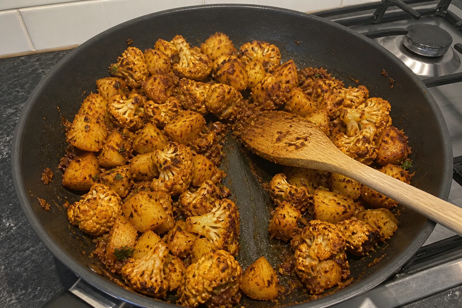 Dry aloo gobi masala clinging to browned potato and cauliflower with a clear spoon track in the pan