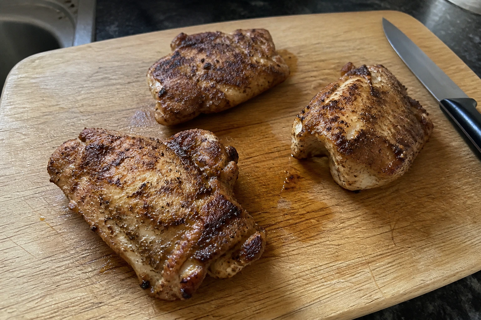 Three unevenly browned chicken thighs resting on a scratched chopping board before slicing